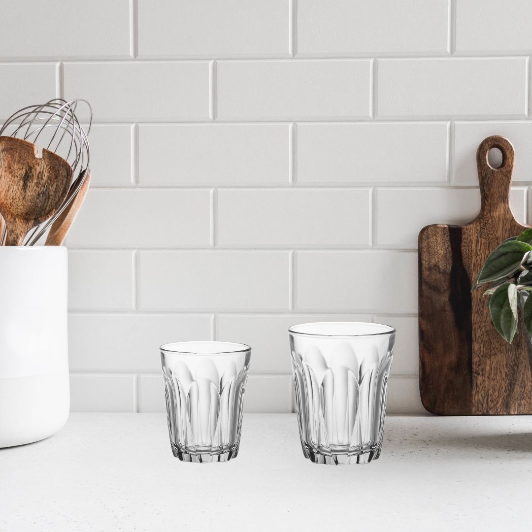 Two clear glasses on a kitchen counter with a white tiled wall in the background.