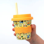 Yellow tumbler with orange lid and straw, held by a hand, on a white background