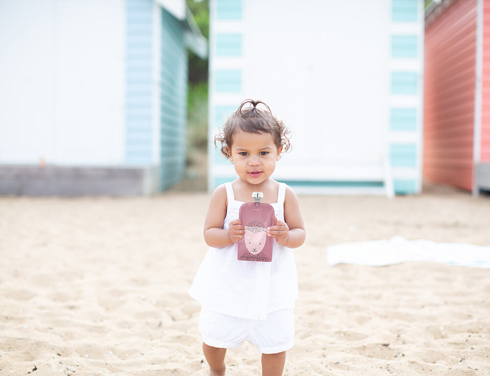 Child in a white dress holding a pink bottle on a sandy beach with colorful huts in the background