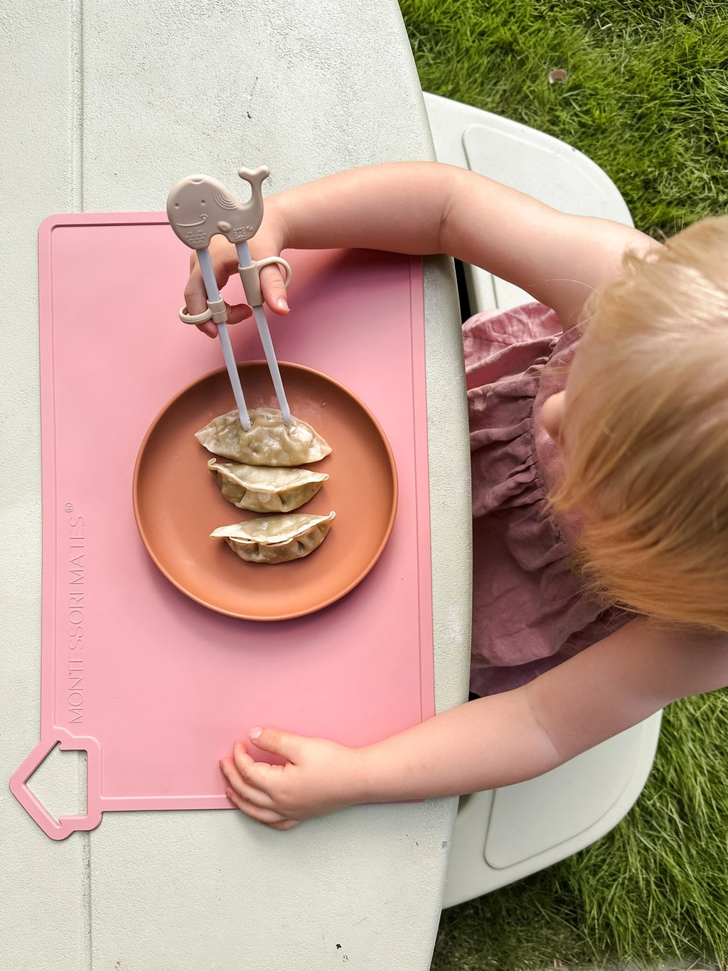 Child using chopsticks to eat dumplings on a pink mat outdoors