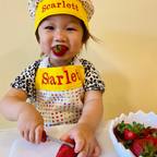 Child wearing a yellow apron and hat with 'Scarlett' on it, eating strawberries.