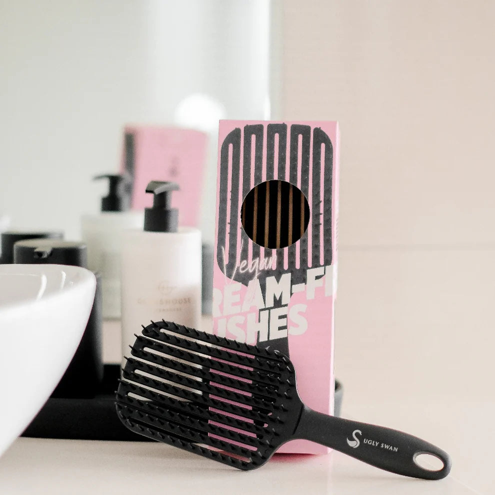 Black paddle brush on a bathroom counter with a pink box labeled 'Scream Free' in the background.