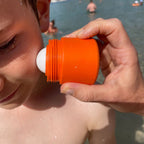 Person applying sunscreen to a child's face with an orange and white container at a beach.
