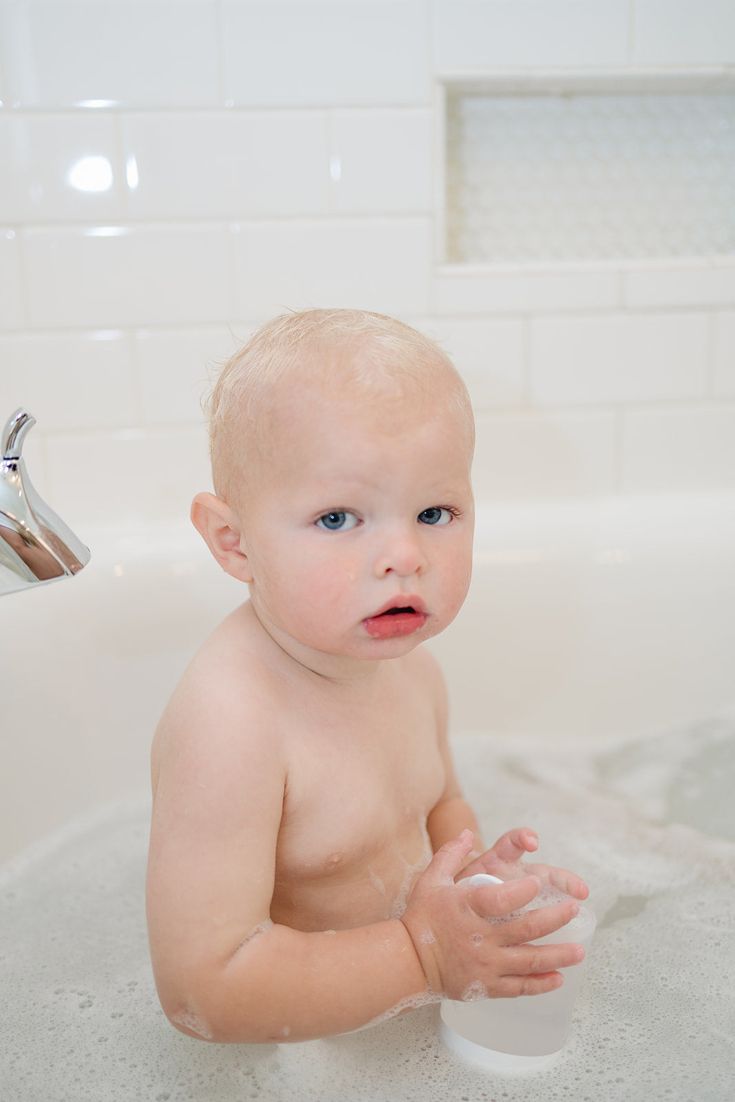 Baby sitting in a bathtub with bubbles, looking surprised or confused.