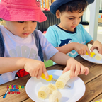 Two children using Safety First Food Kutter to cut bananas at a wooden table outdoors.