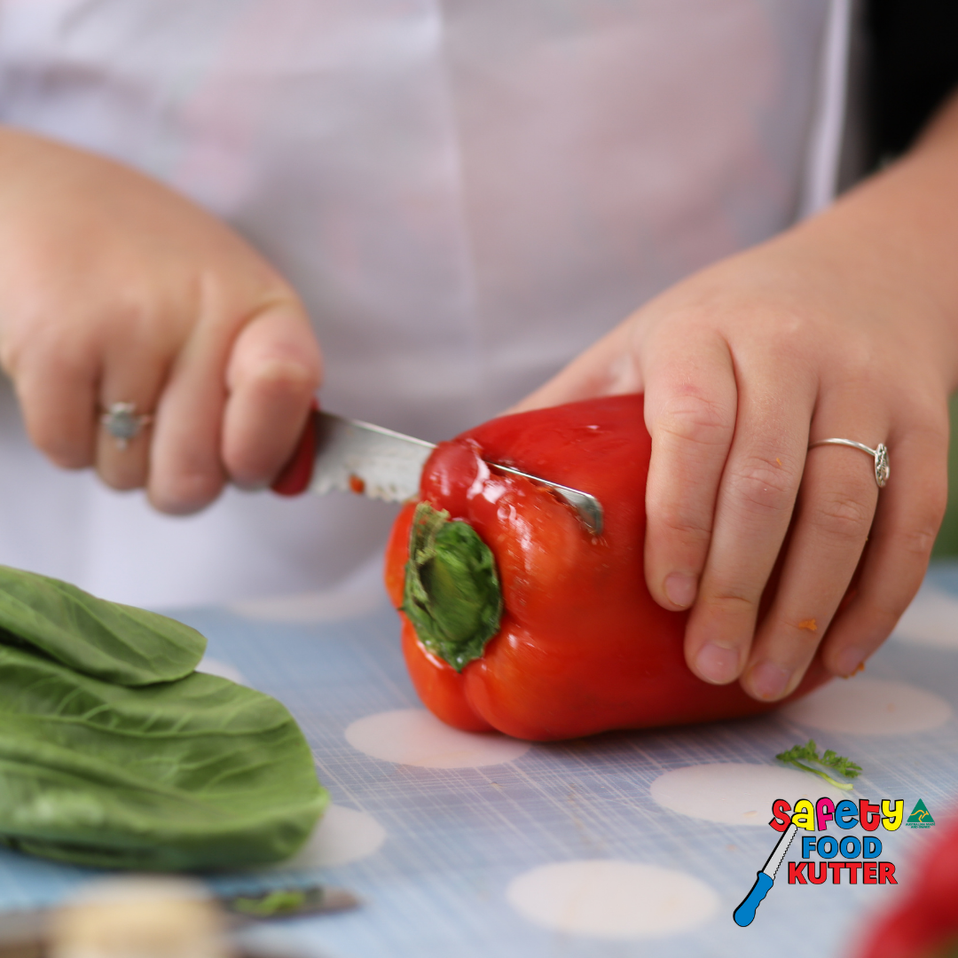 Person cutting a red bell pepper with a knife on a cutting board, featuring the Safety 1st Food Kutter logo.