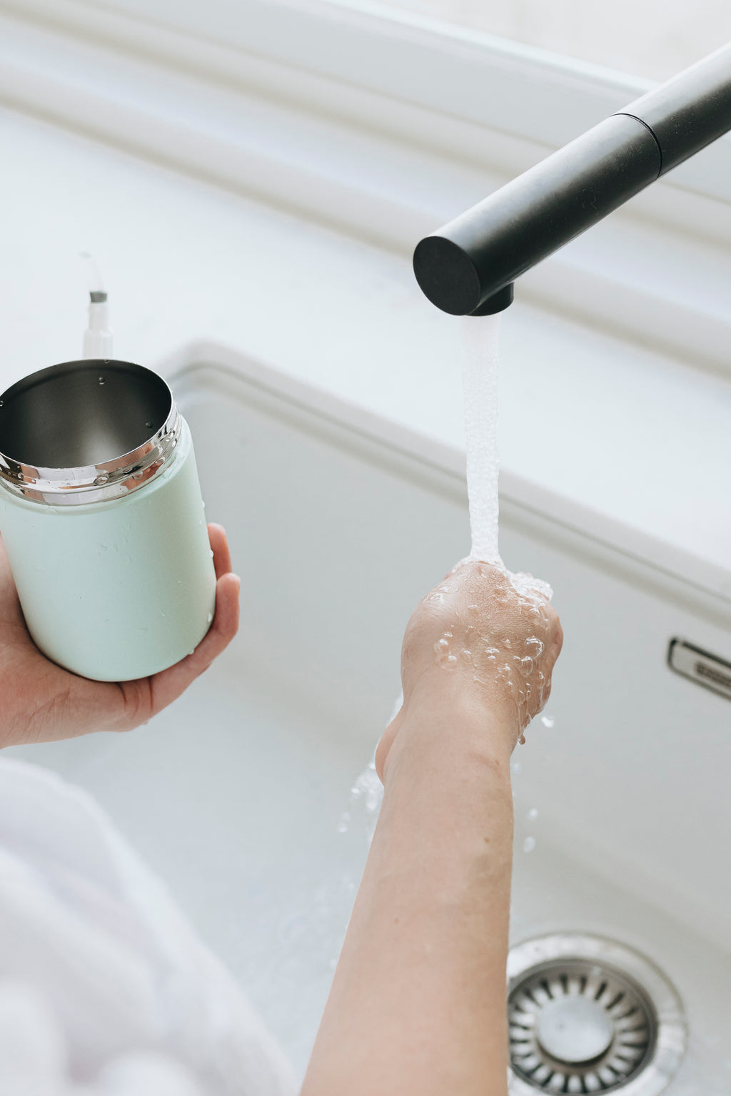 Person washing a wipes dispenser under running water in a kitchen sink.