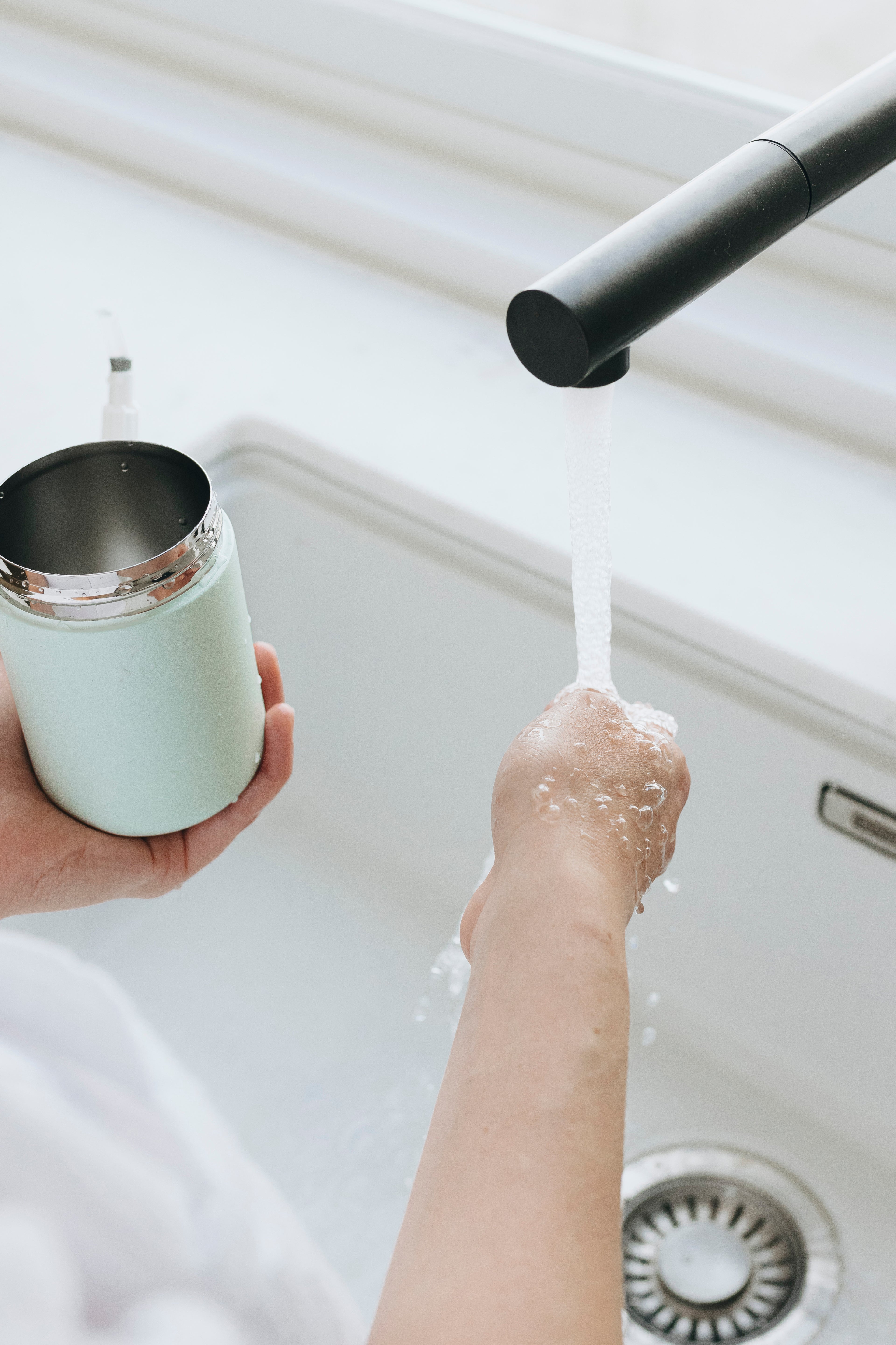 Person washing a wipes dispenser under running water in a kitchen sink.