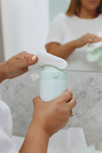 Person holding a water dispenser with a pump, possibly a skincare product, in a bathroom setting.