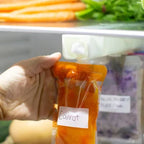 Hand holding a food pouch labelled carrot in front of a container with more carrots and vegetables. The breast milk/food pouch is hanging on the organiser.