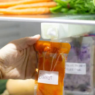 Hand holding a food pouch labelled carrot in front of a container with more carrots and vegetables. The breast milk/food pouch is hanging on the organiser.